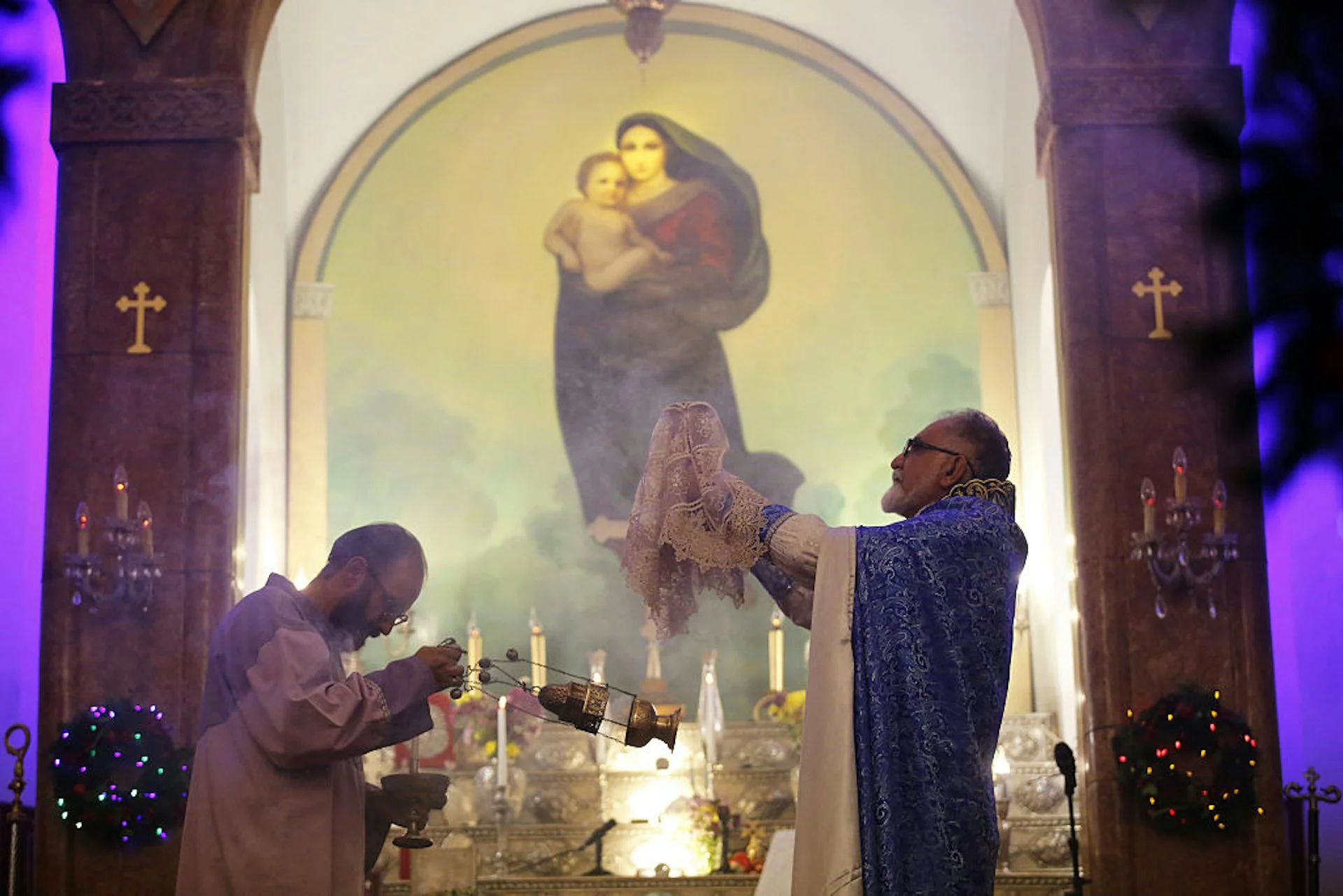 A man in an ornate blue robe holds up an item covered in lace as he stands in front of a mural of Mary and the infant Jesus.