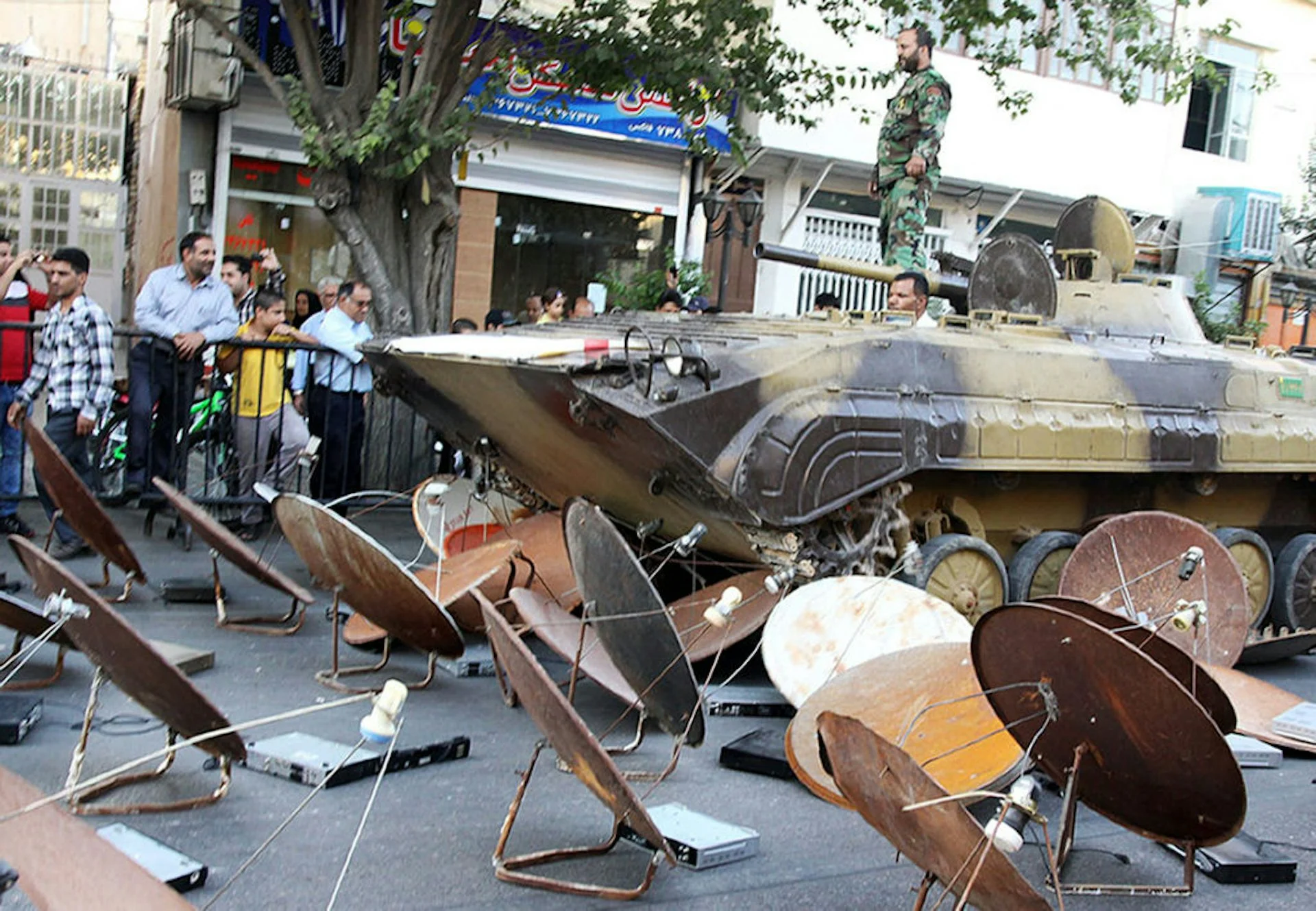 A street full of satellite dishes, with a camouflage-colored tank nearby and shops lining the street.