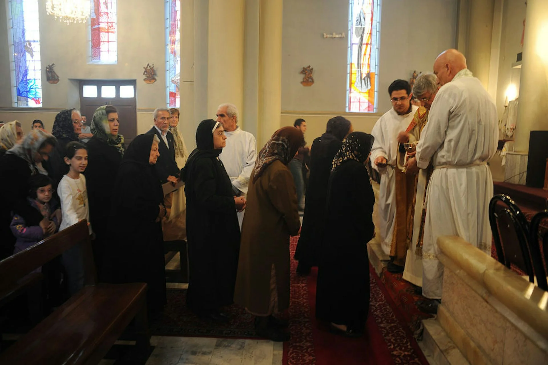 A line of people in dark clothing stands in the aisle of a church, leading up to a few clergymen in white robes.
