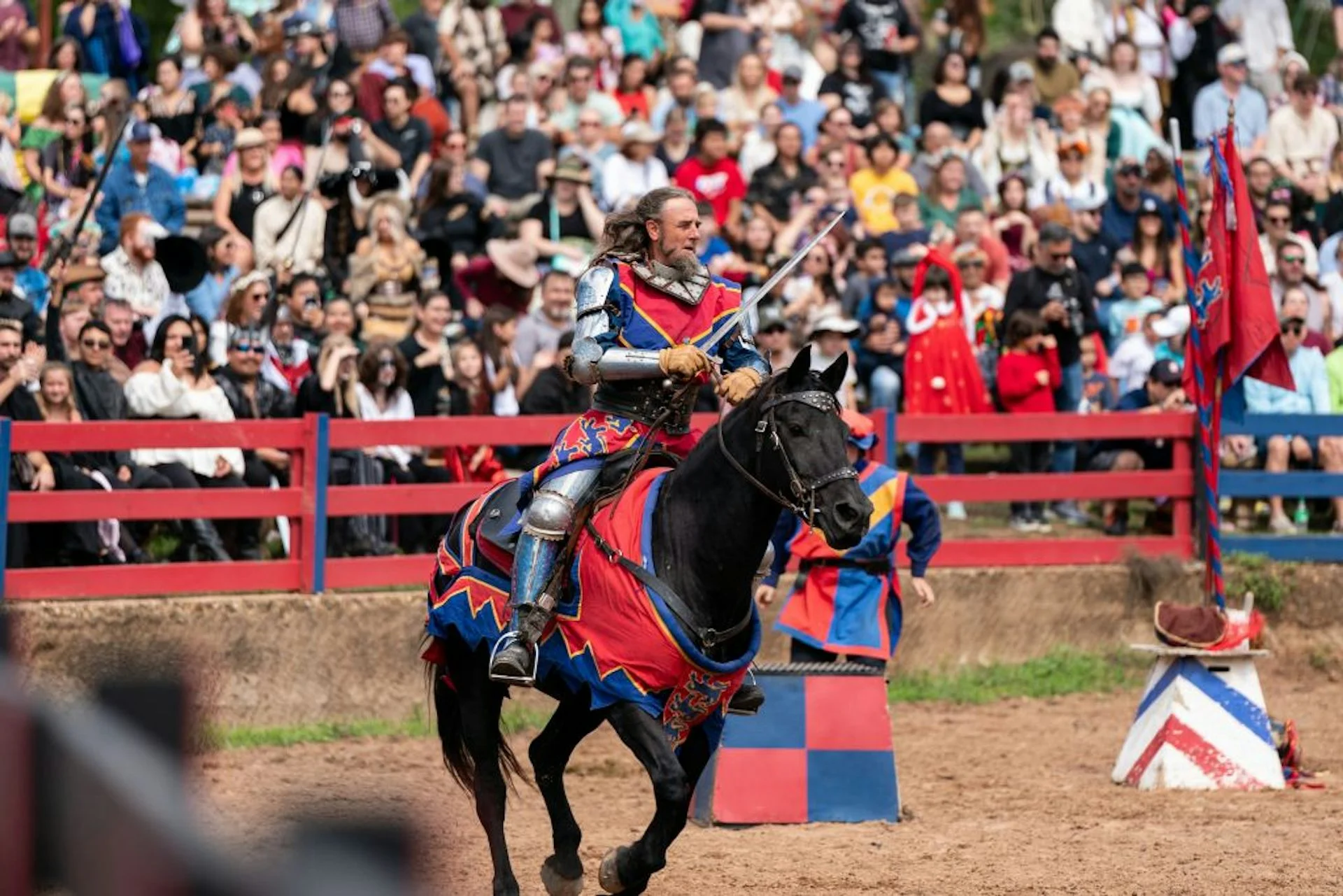 A man rides a horse while holding a jousting lance in front of bleachers full of spectators.