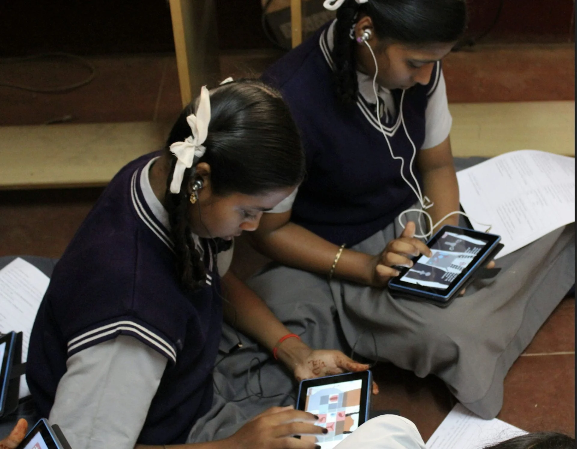 Two girls in a school uniform sit on the floor playing a game on a digital tablet.