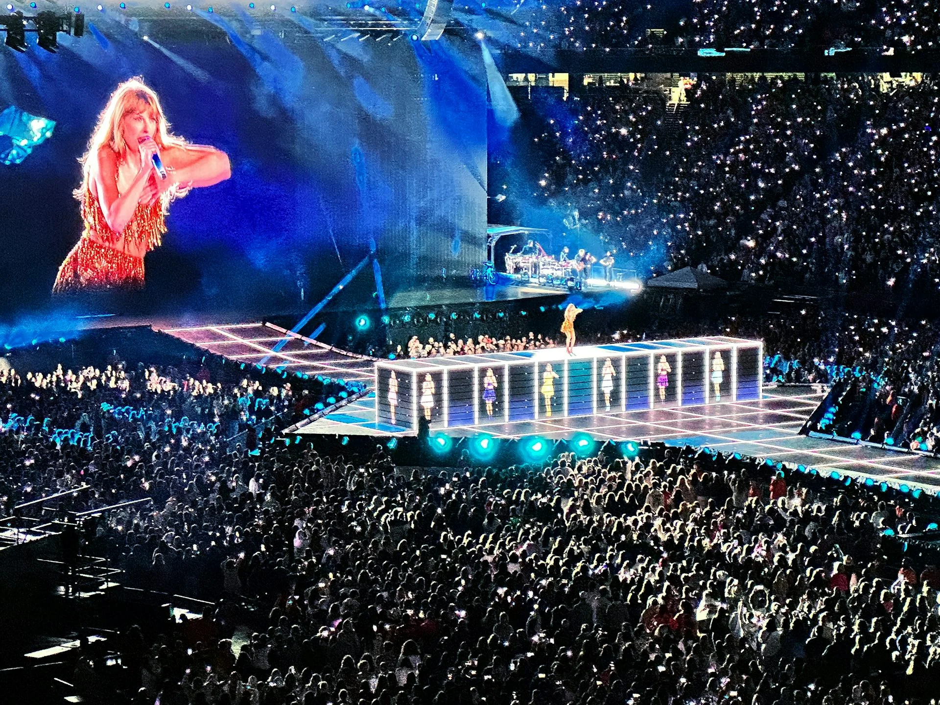 A giant image of Taylor Swift on a screen looms over a concert crowd while she performs on stage during her Eras tour in Foxborough, Mass.