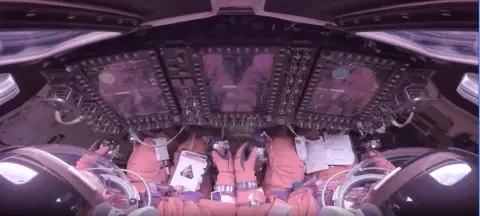 NASA This image looks forward over the shoulders of two Artemis II astronauts strapped into their seats inside the Orion crew module. They wear bulky orange pressure suits and clear helmet bubbles, facing a wall of tall, glowing display screens and dense rows of switches and knobs. Their gloved hands rest near hand controllers and a checklist. Around the edges of the frame, curved windows, cables and equipment make the cabin look compact and highly technical