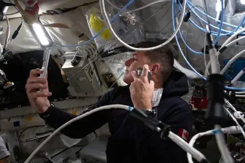 NASA This close interior image shows Canadian astronaut Jeremy Hansen floating inside the cramped Orion spacecraft while shaving. He wears a dark hooded top with a small Canadian flag patch on the sleeve and holds a razor up to his cheek. Around him, the cabin walls are lined with white padded panels, storage bags and equipment boxes. Blue and white cables loop across the frame, and bright strip lighting overhead makes the small space look crowded, practical and lived in.