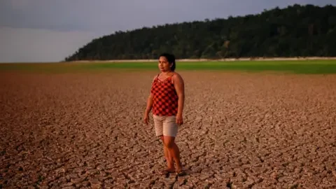 Reuters A woman in a red and black vest top, beige shorts and sandals stands on a dried-up river bed. There is a forest in the background.