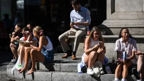 Getty Images Men and women sit on the steps of a fountain in the sunshine while eating their lunch and checking their phones. A number of women are wearing shorts. 