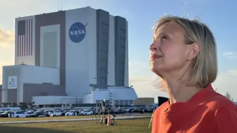 BBC/Kevin Church BBC Science Editor Rebecca Morelle stands about 100 metres in front of the cuboid Nasa mission control building. She is looking into the Sun as a shadow falls across the grass between her and the building. Some camera tripods are visible in the middle ground and a car park. Rebecca wears a red blouse or dress.