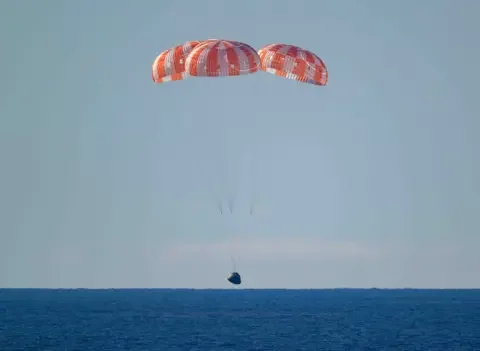 NASA The landing craft with it's orange parachutes lands on a dark blue sea.