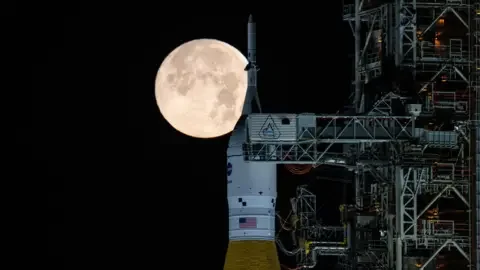 NASA A large and very bright full Moon appears directly behind the top of Nasa's Space Launch System rocket at night. The orange and white rocket is sitting next to a steel support structure called a mobile launcher.